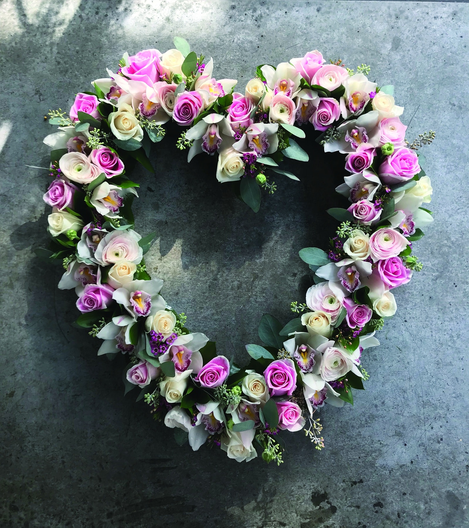 A delicate pink and white open heart made with pink and white roses, blush rununculus, and white cymbidium orchids nestled in with seeded eucalyptus and ruscus.