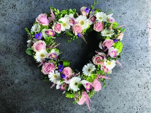 An open heart design with green hydrangea, soft pink roses, white cymbidium orchids, soft pink astilbe, white mini gerberas, purple freesia, and purple limonium nestled among eucalyptus.