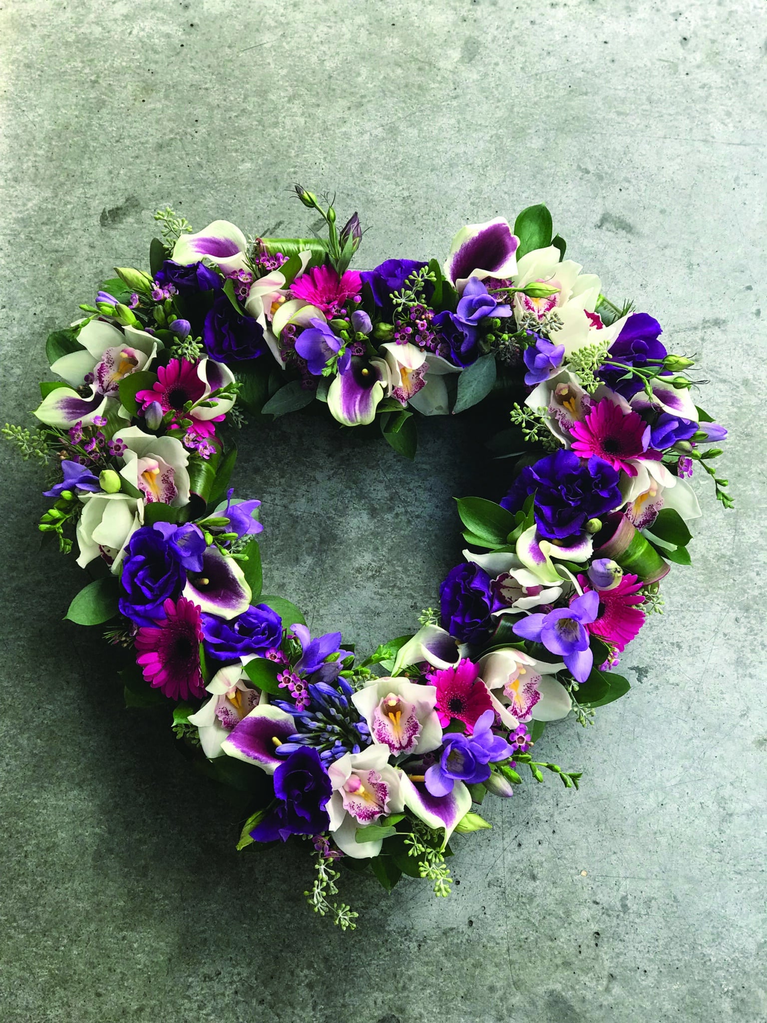 A pink and purple open heart design made with calla lilies, cymbidium orchids, agapanthus, purple freesia, pink gerbera daisies, puprle lisianthus, and pink waxflowers.