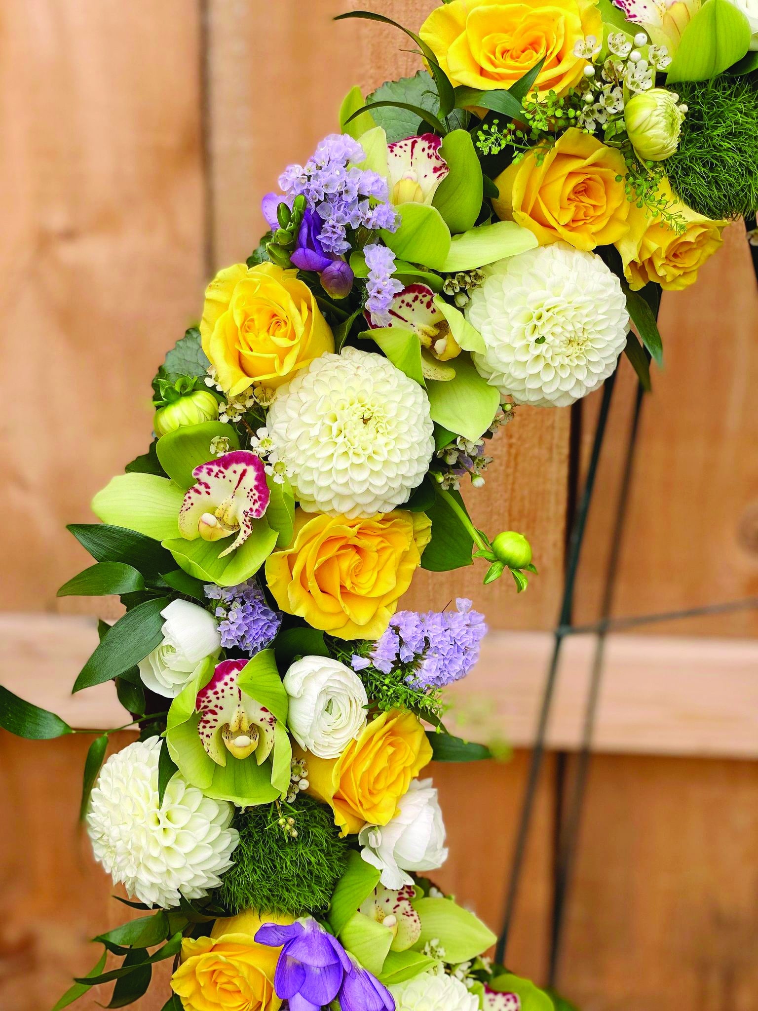Close up of the white dahlias, yellow roses, green cymbidium orchids, purple freesia, and blue statice on a floral funeral wreath.