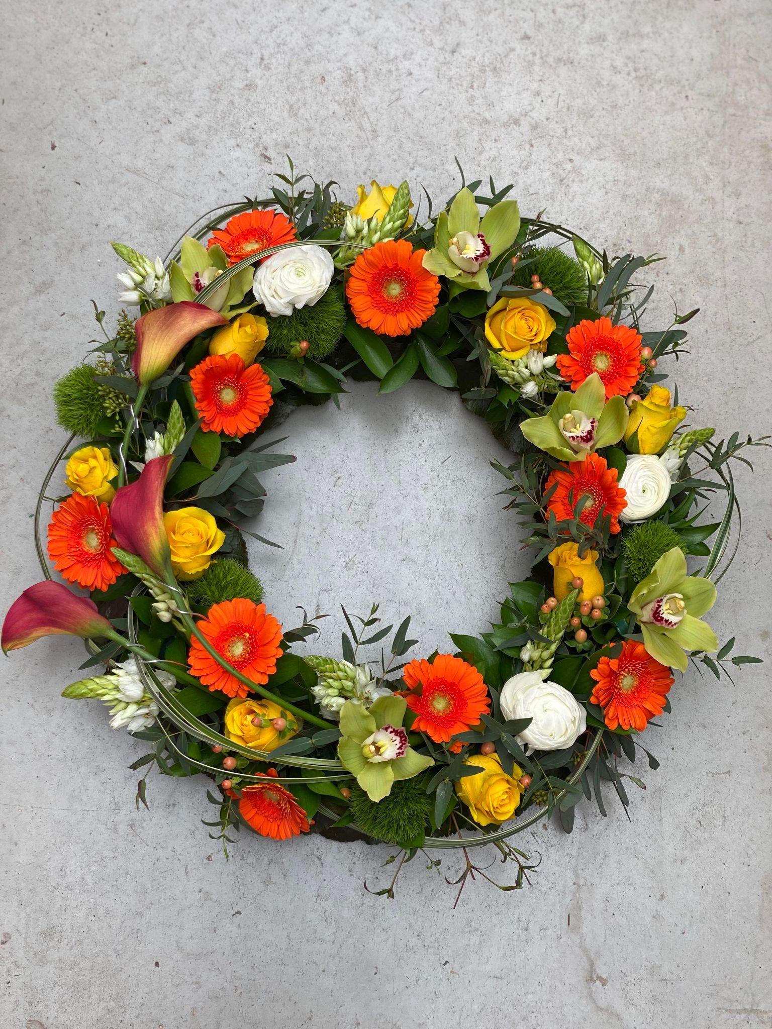 A medium bevreavement wreath made with orange calla lilies, orange gerbera daisies, yellow roses, green cymbidium orchids, white ranunculus, peach hypericum, and star of Bethlehem on an abundant bed of greenery.