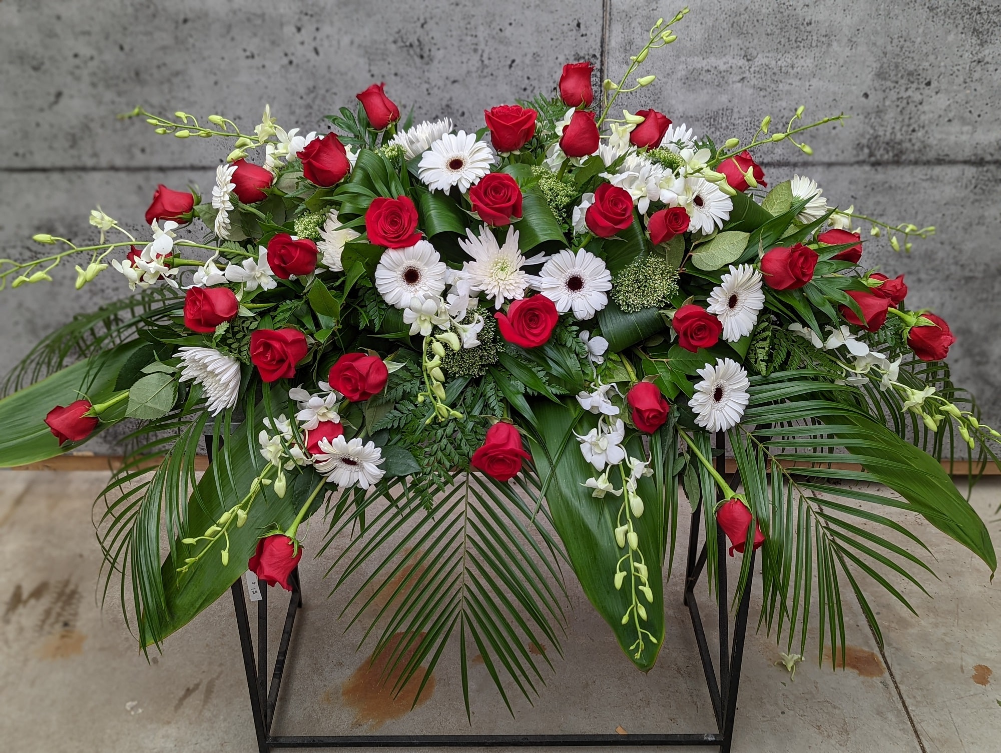 A red and white casket spray made with red roses, white mums, white trachelium, white mini gerbera daisies, and white dendrobium orchids in a bed of lush greens.