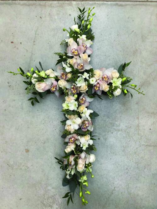 A soft pink and white flowered cross featuring cymbidium orchids, debdrobium rochids, roses, and ranunuculus.