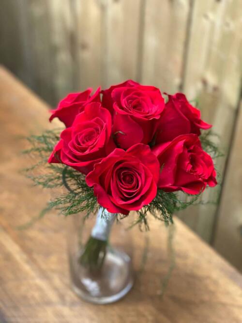 a bouquet of 7 red roses with delicate tiki fern around the outside and white ribbon wrapped around the stems in a glass vase.