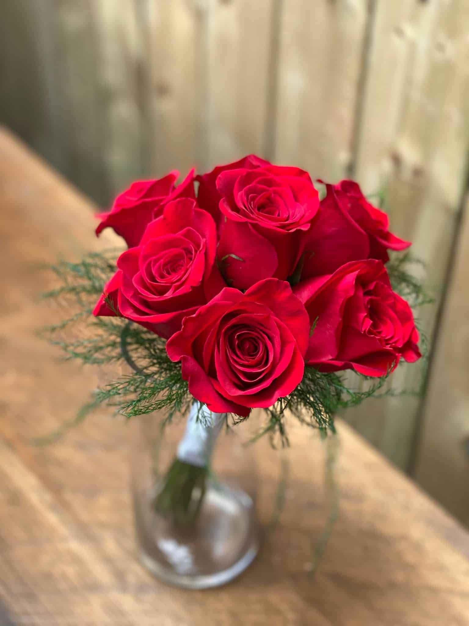 a bouquet of 7 red roses with delicate tiki fern around the outside and white ribbon wrapped around the stems in a glass vase.
