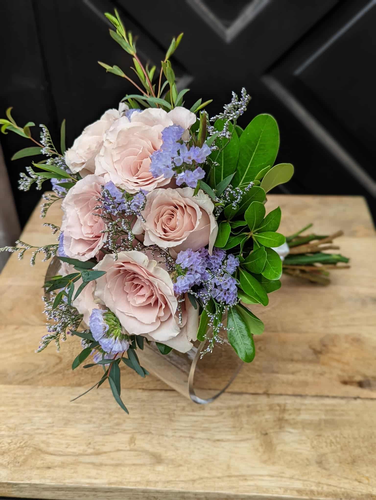 A bouquet of blush roses, blue statice, limonium, and soem greenery satin wrapped lying on a table.