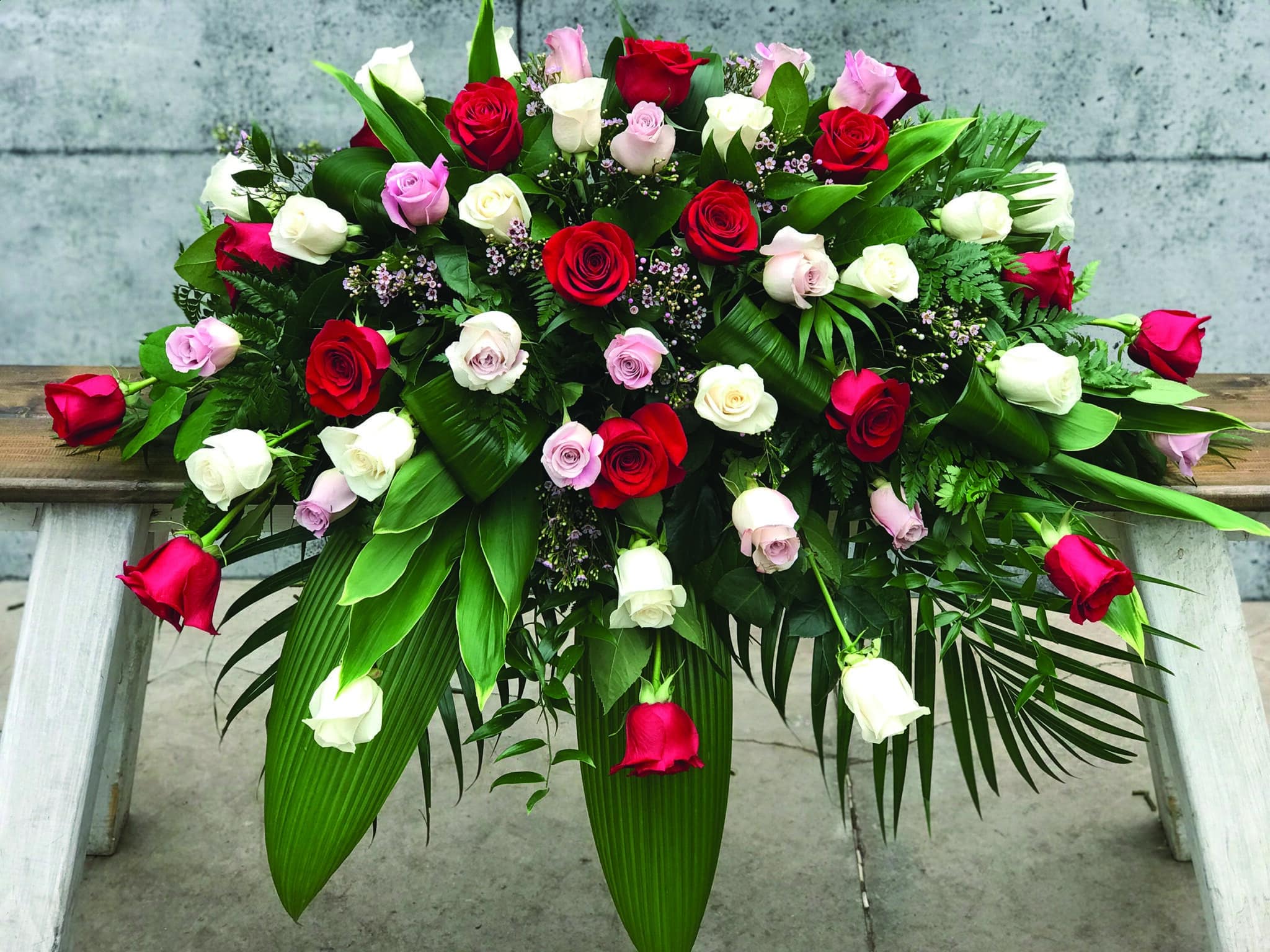A casket spray made with an assortment of red, white, and pink roses, supported by waxflower and a variety of greens.