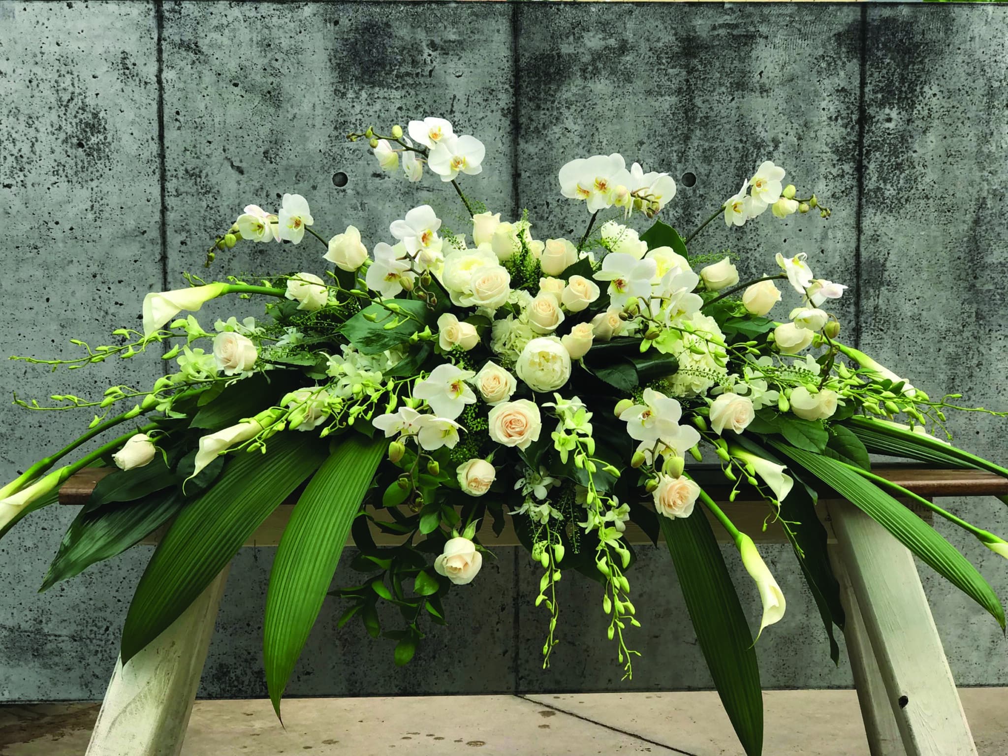 An all white casket spray made with calla lilies, dendrobium orchids, hydrangea, peonies, roses, and phalaenopsis orchids in a bed of tropical greens.