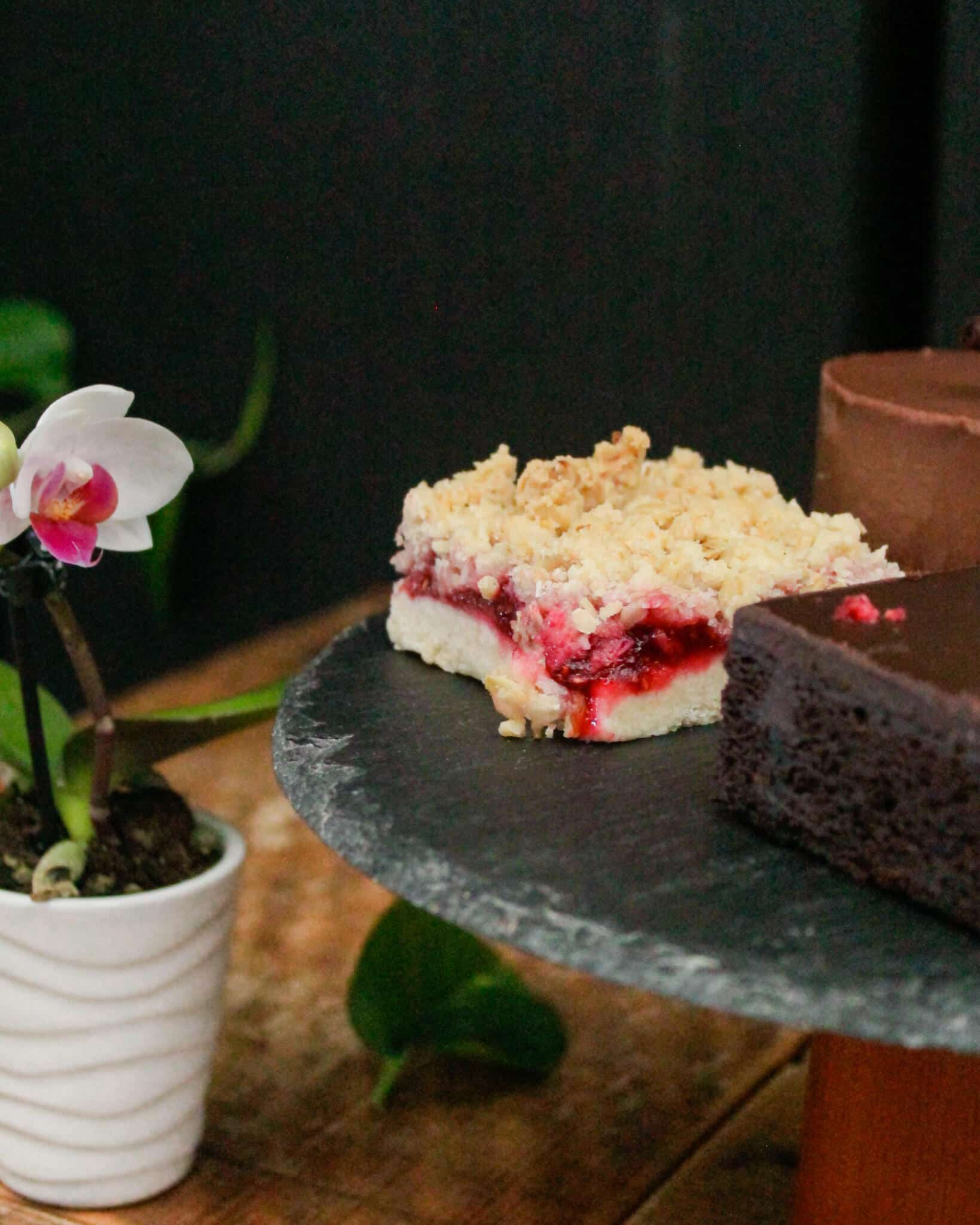 A close-up of a raspberry crumble bar and a chocolate brownie on a slate stand, with a pink orchid in a white pot.