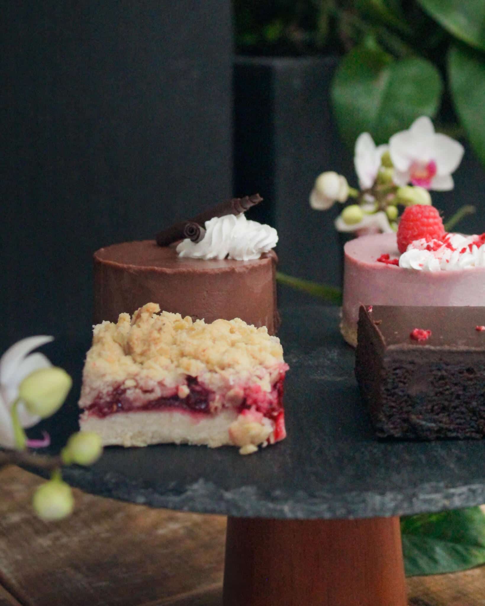 A slate cake stand with a chocolate mousse mini cake, a raspberry crumble bar, a raspberry mousse cake topped with a fresh raspberry, and a chocolate brownie, garnished with a white orchid.