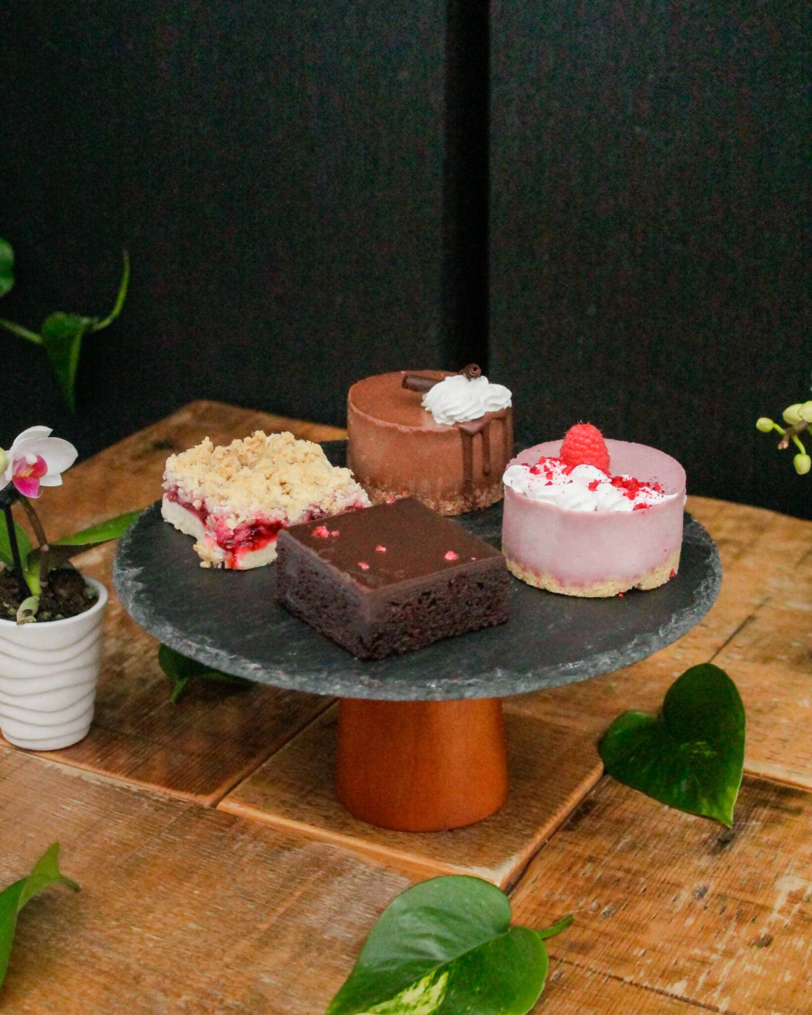 A slate cake stand with a raspberry crumble bar, a chocolate mousse mini cake with whipped topping, a chocolate brownie, and a raspberry mousse cake topped with fresh fruit, with an orchid.