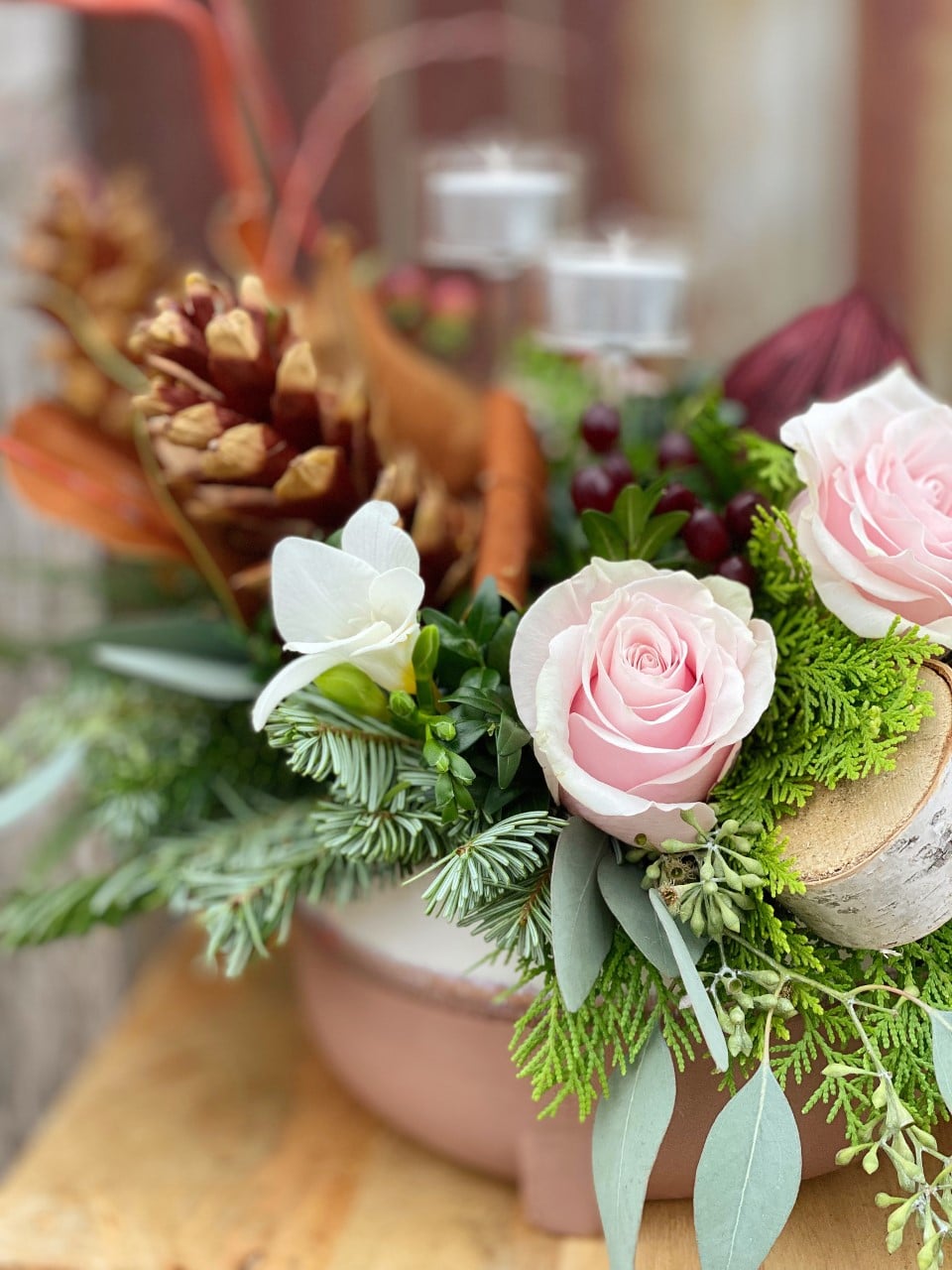 A close-up of a winter centrepiece with pink roses, white freesia, red berries, pinecones and cedar in a blush oblong pot.