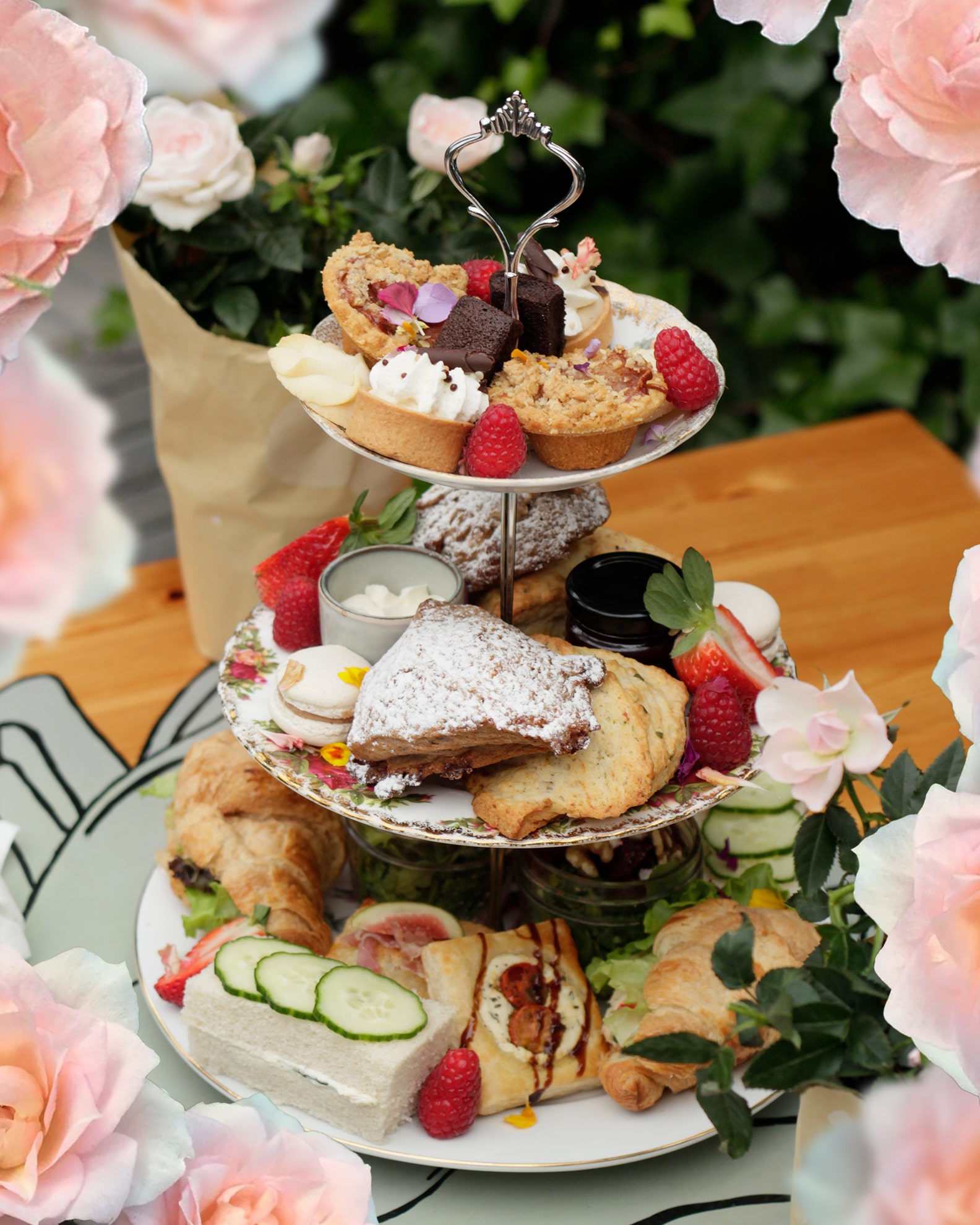 A three-tier stand filled with pastries, scones, tarts and finger sandwiches with fresh berries, cream and jam, set among pink roses for the Garden Tea.