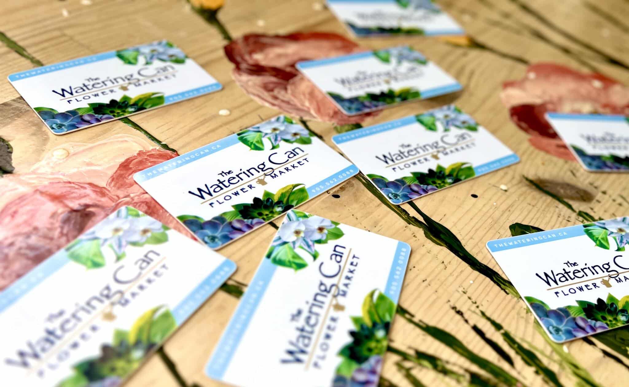 Watering Can Gift Cards The Watering Can Flower Market gift cards scattered across a hand-painted floral table.