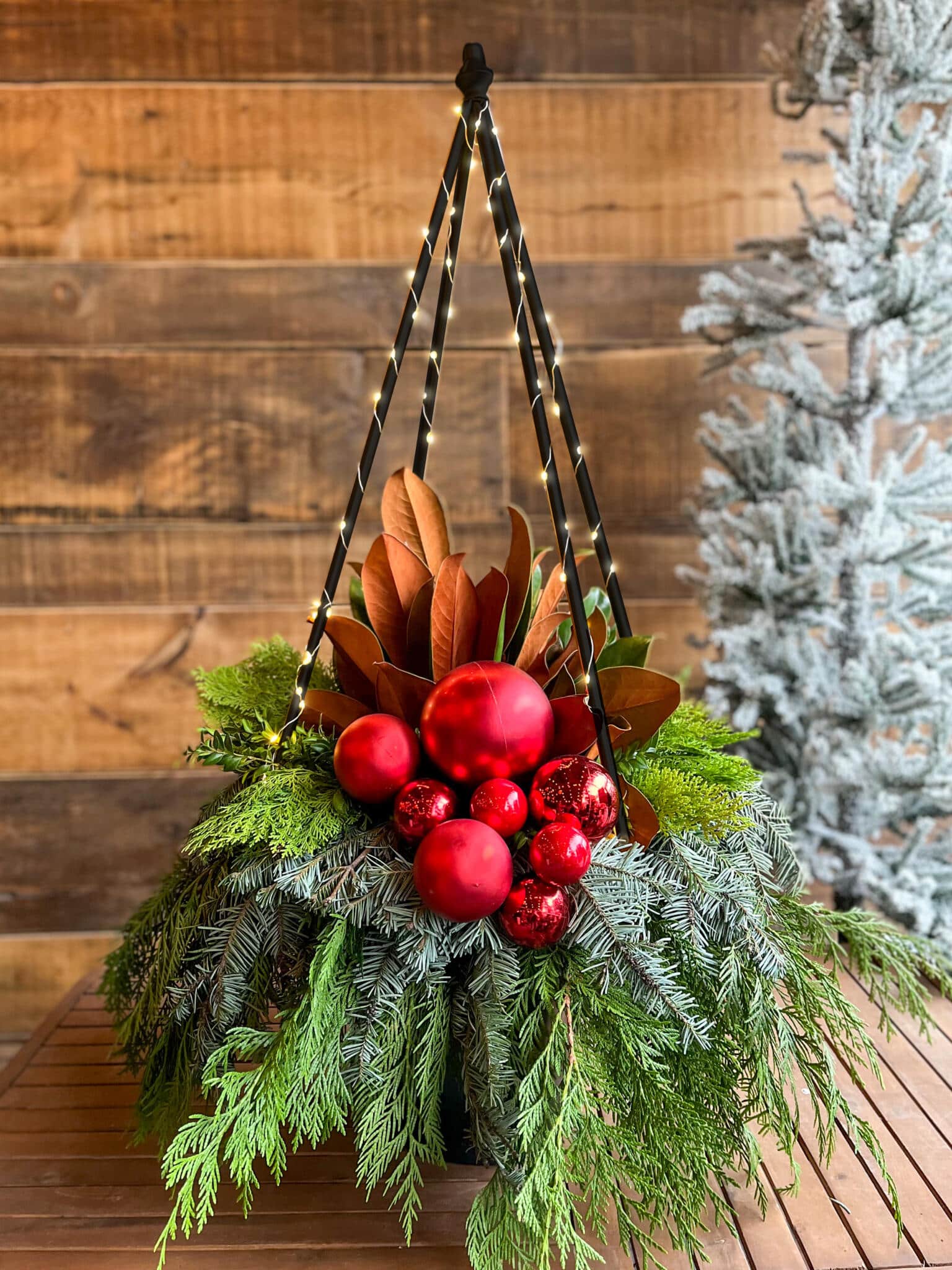 A large black metal tree/pyramid with lights on abed of winter greenery, magnolia and red ornaments