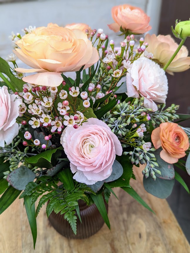 The Peach Dreams arrangement of peach roses, pink ranunculus, and wax flower surrounded by soft greenery.