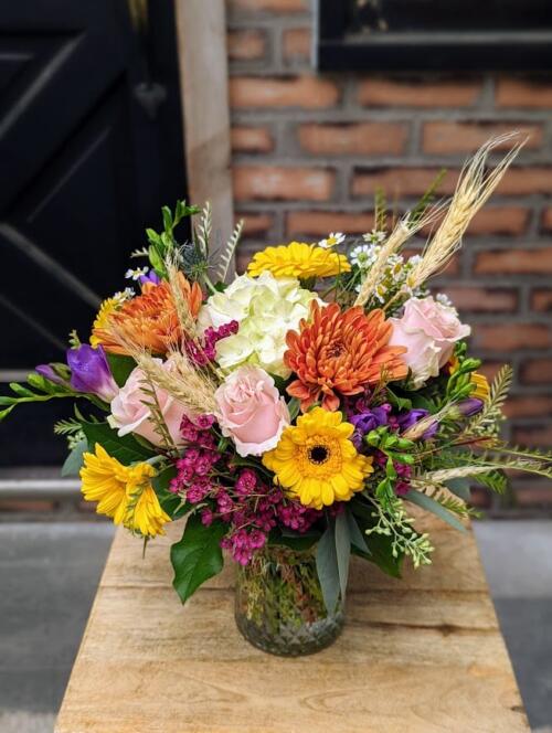 A colourful autumn bouquet in a glass vase with yellow gerbera daisies, an orange chrysanthemum, pink roses, white hydrangea and wheat.