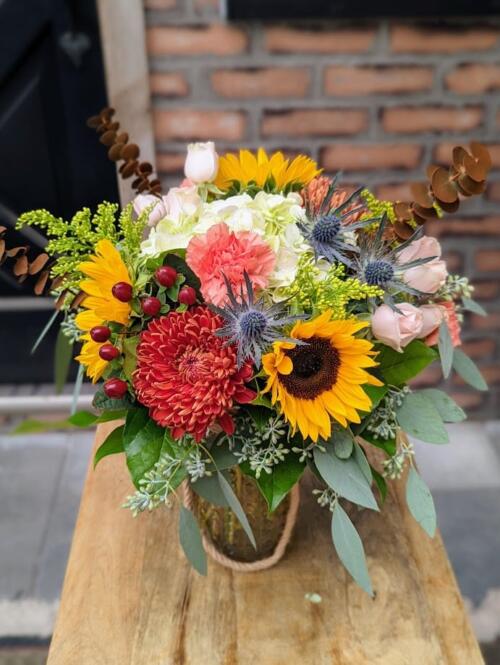 An amber vase with a rope handle overflowing with fall toned blooms. featuring rust coloured mums, yellow sunflowers, thistle, peach carnations, brown eucalyptus and a large antique hydrangea.