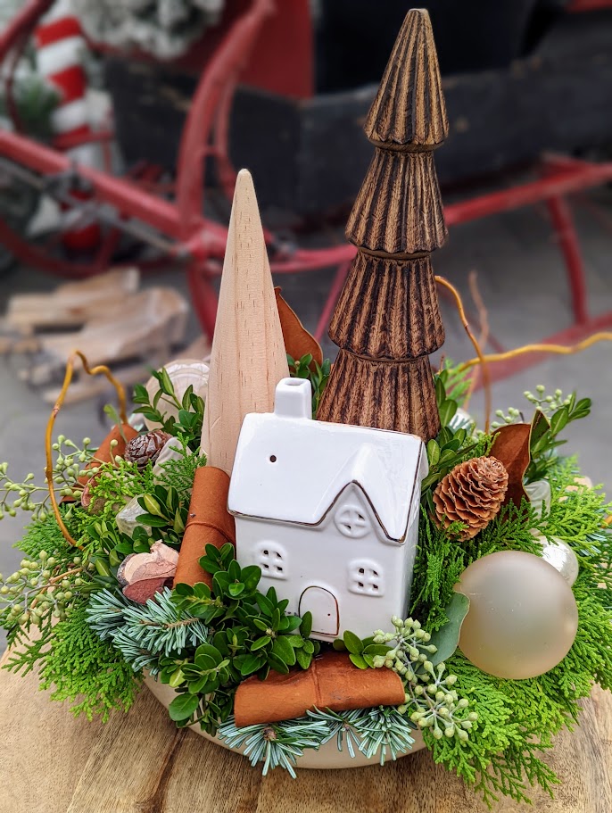 A festive tabletop arrangement in a bowl with wooden tree figures, a white ceramic house, fresh cedar and boxwood, cinnamon sticks, pinecones, and a gold ornament.