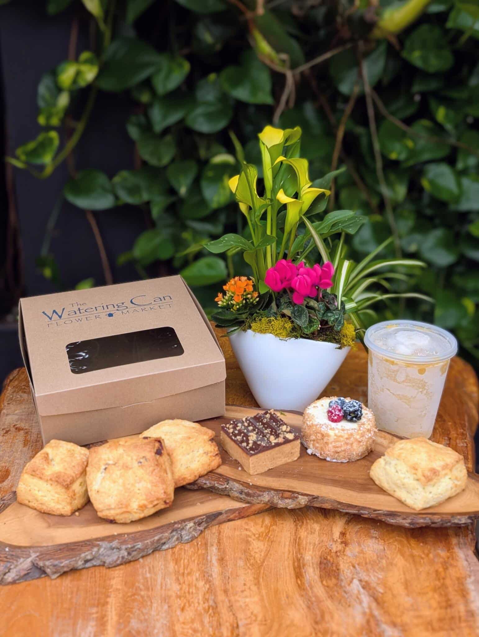 The Cheer Collection from The Watering Can displayed outdoors on a wooden table, with a green gift box, house-made treats, drinks, and a small plant arrangement.