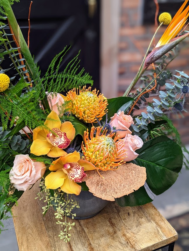 The Aloha Blooms arrangement featuring a yellow cymbidium orchid, an orange pincushion protea, roses, and tropical foliage.