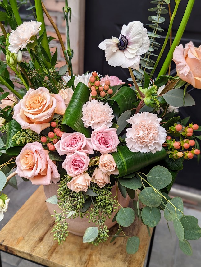 A soft arrangement of blush and peach roses, carnations, a white anemone, and hypericum berries with aspidistra leaves and eucalyptus in a taupe ceramic pot.