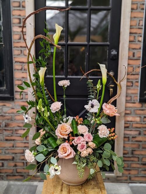 A tall, elegant arrangement of white calla lilies, peach roses, carnations, and anemones with curly willow and eucalyptus in a taupe ceramic pot.