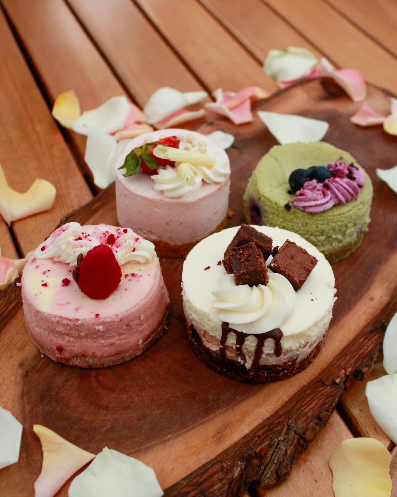 Four handmade mini cakes on a wooden board with rose petals at The Watering Can Pastry Market — including a strawberry mousse, a matcha blueberry, a raspberry mousse, and a chocolate drip mousse cake.