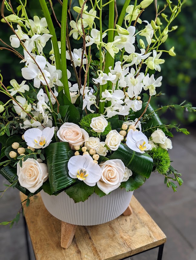 A white floral arrangement of orchids, cream roses, and lush greenery in a white footed pot.