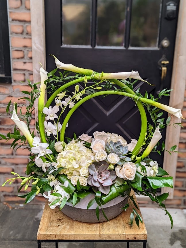 A sculptural arrangement with white calla lilies arching into a hoop above white hydrangea, cream roses, white orchids, and rosette succulents in a stone bowl, set against a black door.