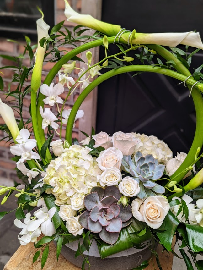An elegant arrangement of white calla lilies arching in a circle above white hydrangea, cream roses, white orchids, and blue-grey and burgundy rosette succulents.