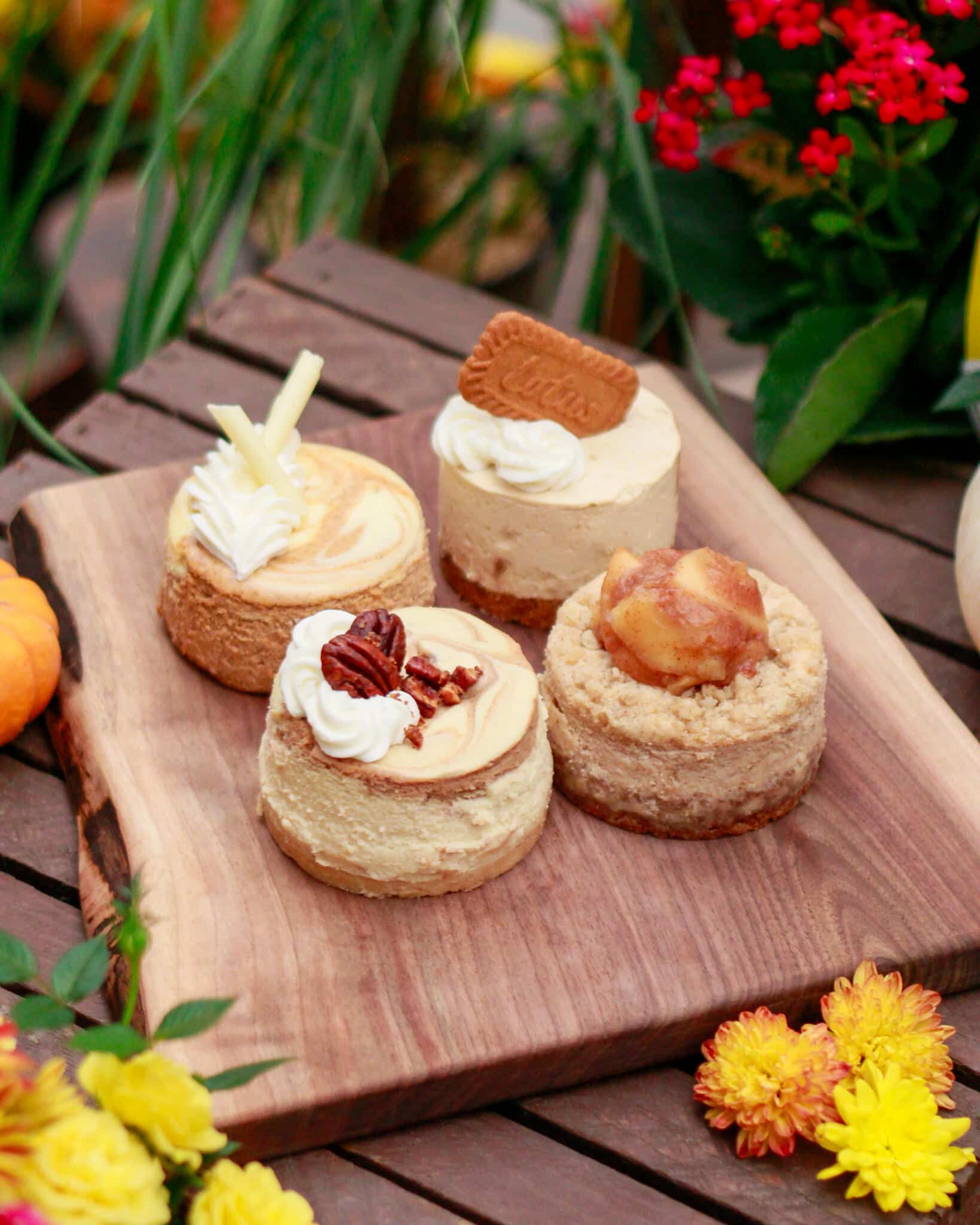 Four mini cheesecakes on a wooden board seen from above at an angle, with red flowers behind and orange mums below.