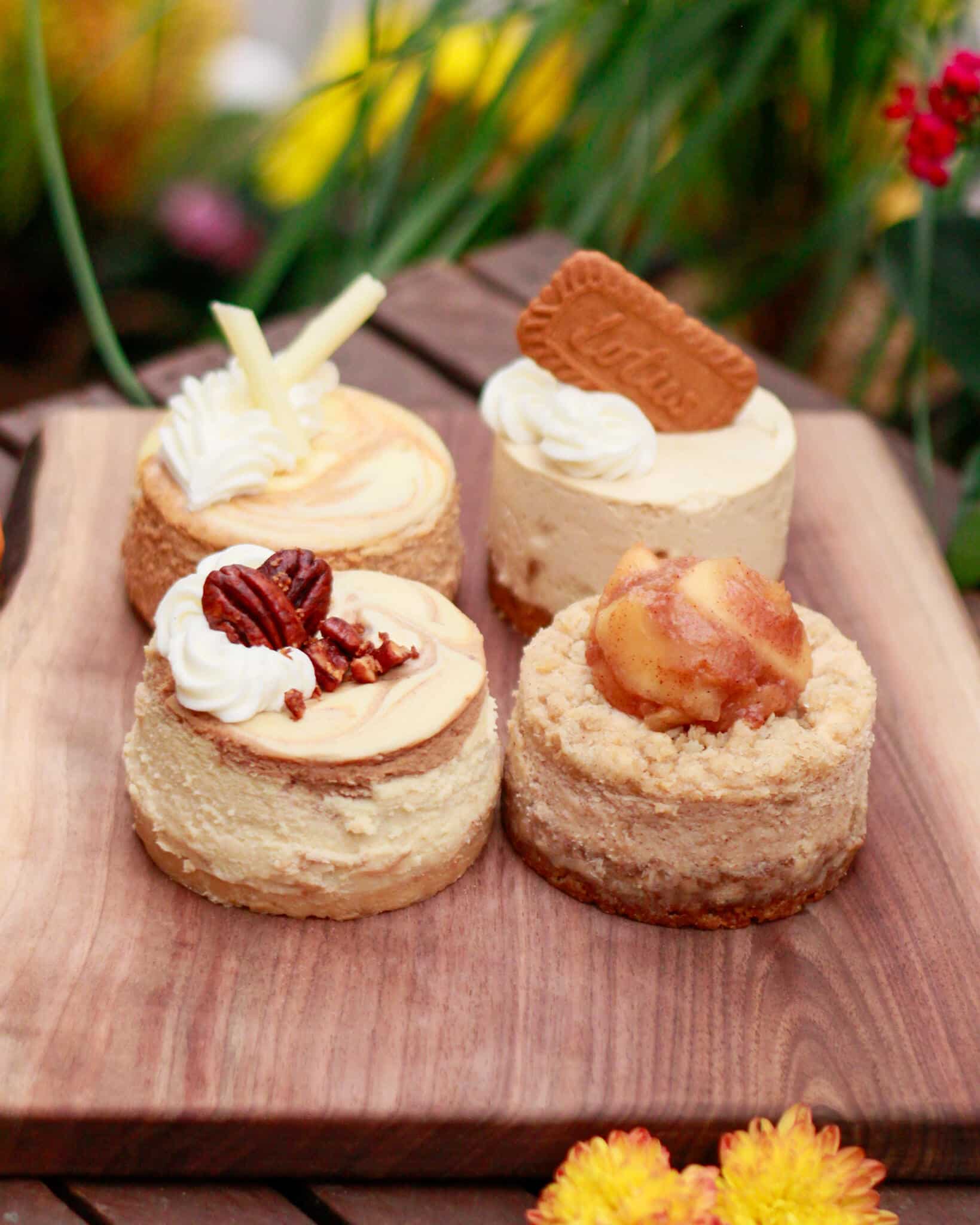 Four mini cheesecakes on a wooden board, angled view, with yellow mums in front and red flowers behind.