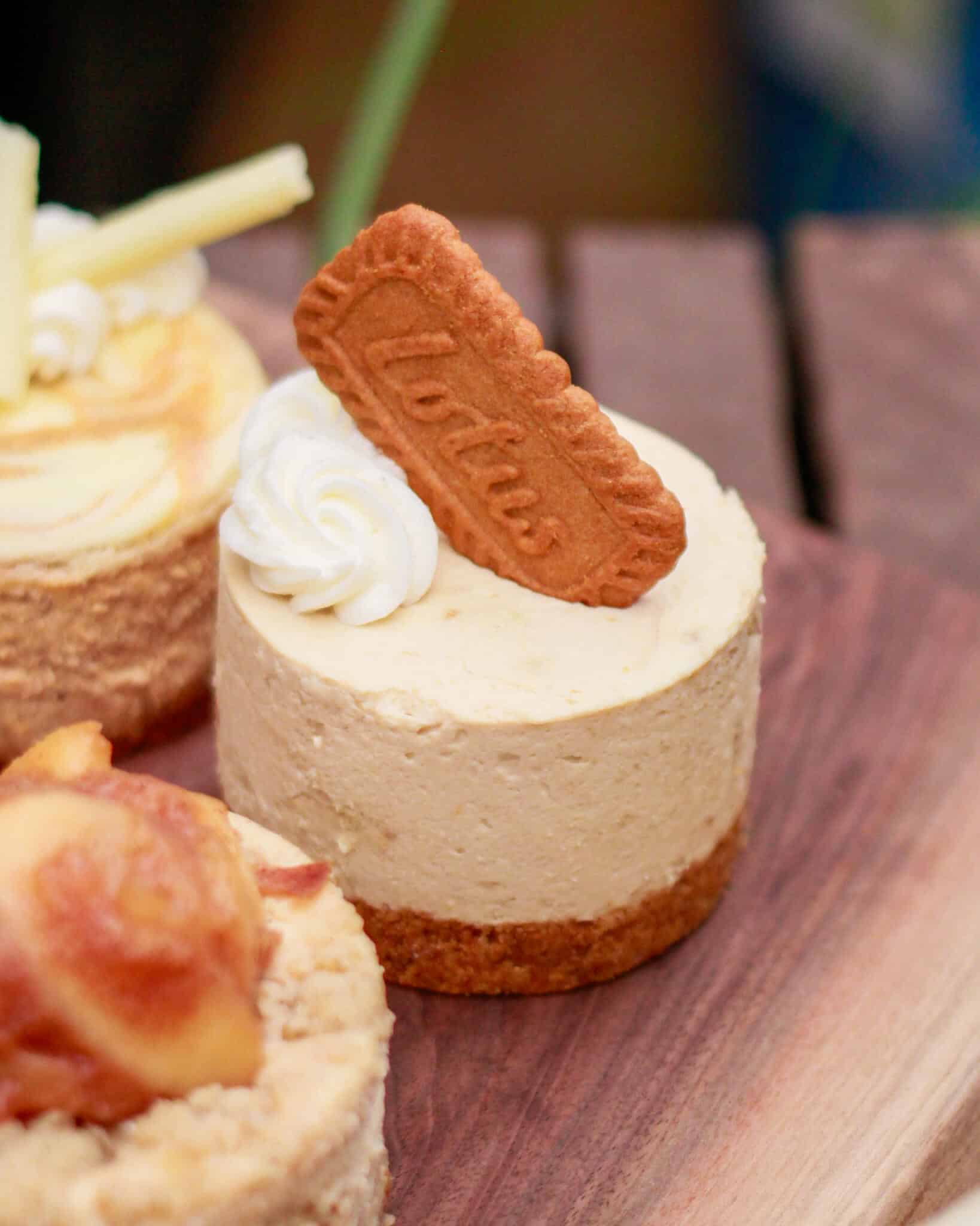 Mini cheesecake topped with a spiced cookie and whipped cream, beside other cheesecakes on a wooden board.