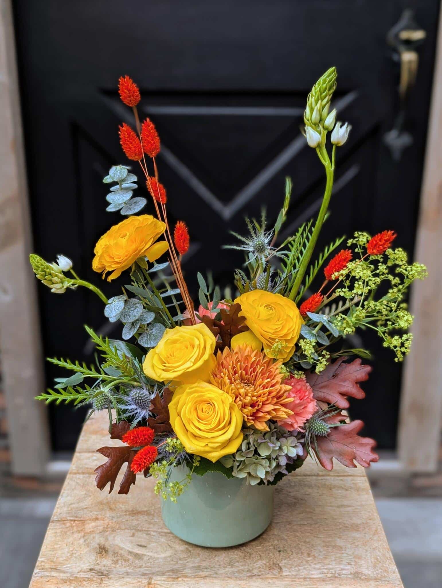 An autumn floral arrangement of sunny yellow ranunculus and roses, a burnt-orange chrysanthemum, red celosia spikes, blue thistle and eucalyptus in a sage-green ceramic vase.