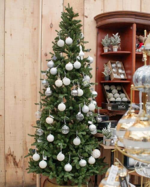 A green Killington Fir artificial Christmas tree trimmed with white and silver ornaments, displayed against a wood-panelled wall in the shop.