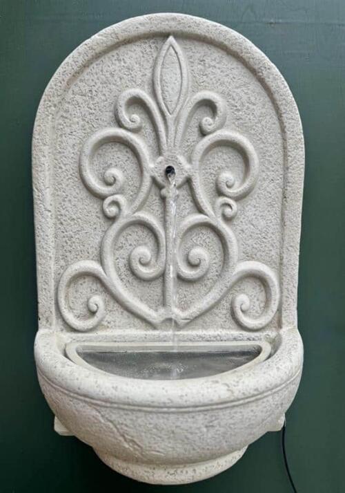 A classic cast-stone wall fountain with an ornate scrollwork back panel above a curved basin.