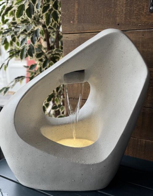 A cream-toned speckled stone-textured sculptural fountain with an organic rounded hoop form and central circular opening, water flowing from a small top spout into a warmly LED-lit pool.