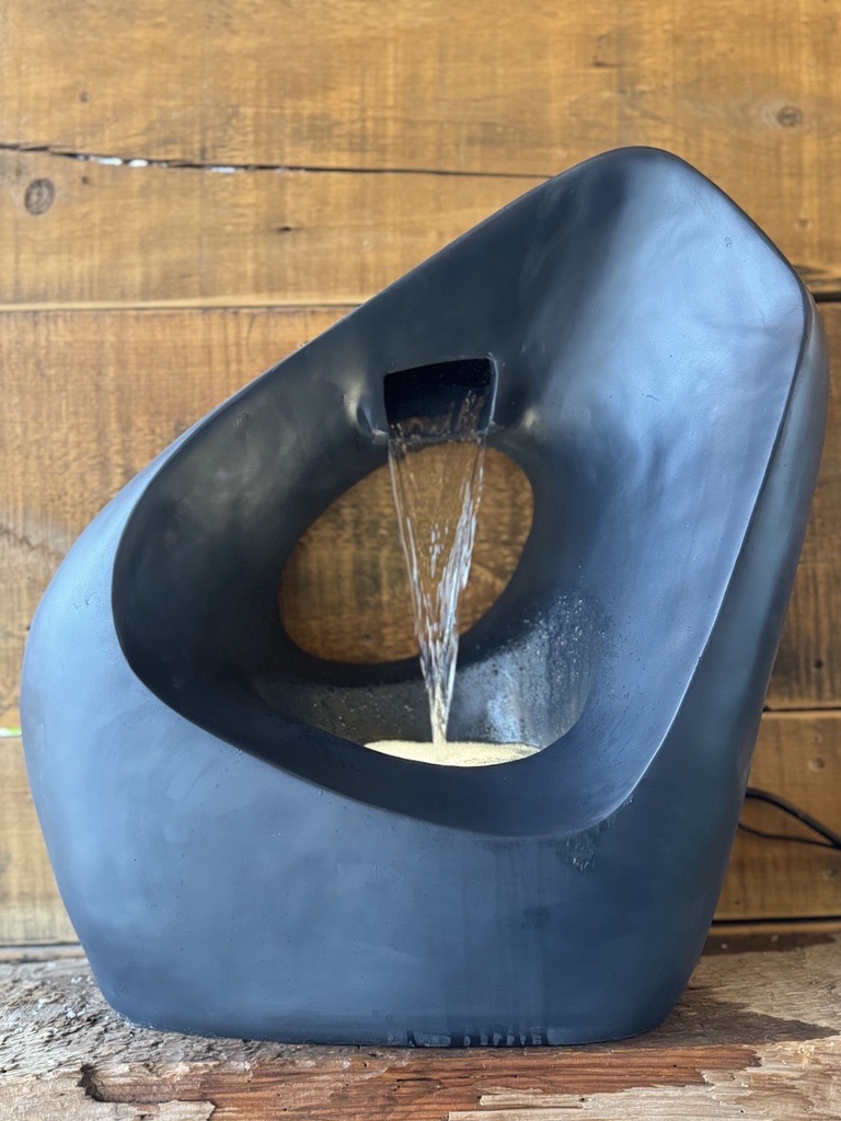 A dark slate-blue sculptural pour fountain with a smooth organic form and central circular opening, water cascading gently from a top spout through the hollow interior.