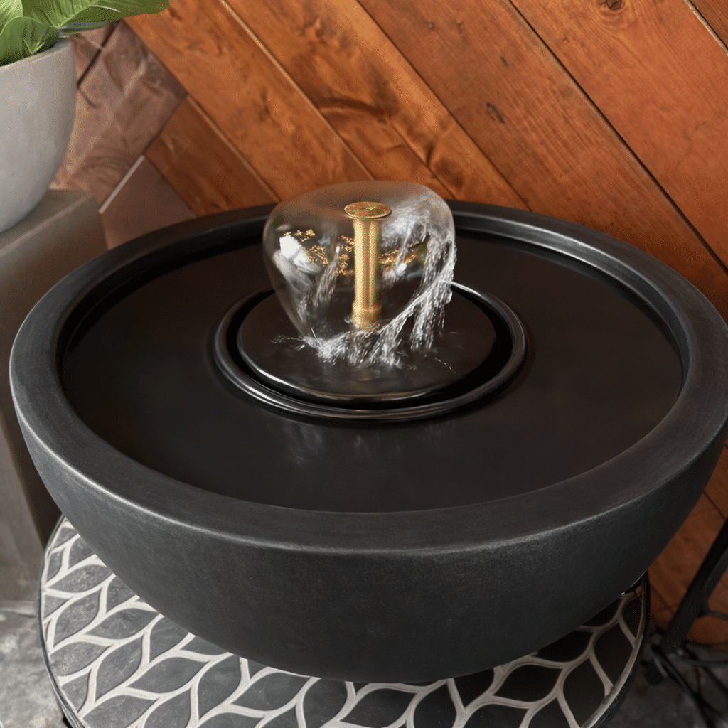 A low round black bowl fountain with a clear bubbling glass orb at its centre, set on a patterned base.