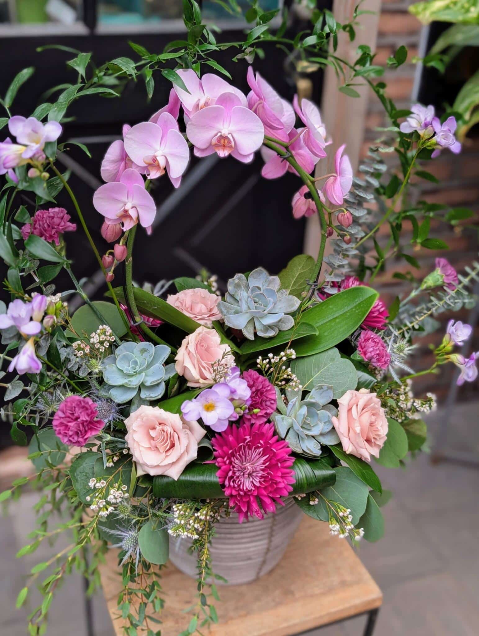 Close-up of an Amethyst Skies floral arrangement featuring purple phalaenopsis orchids, blush roses, magenta chrysanthemums, and succulents in a white ceramic pot