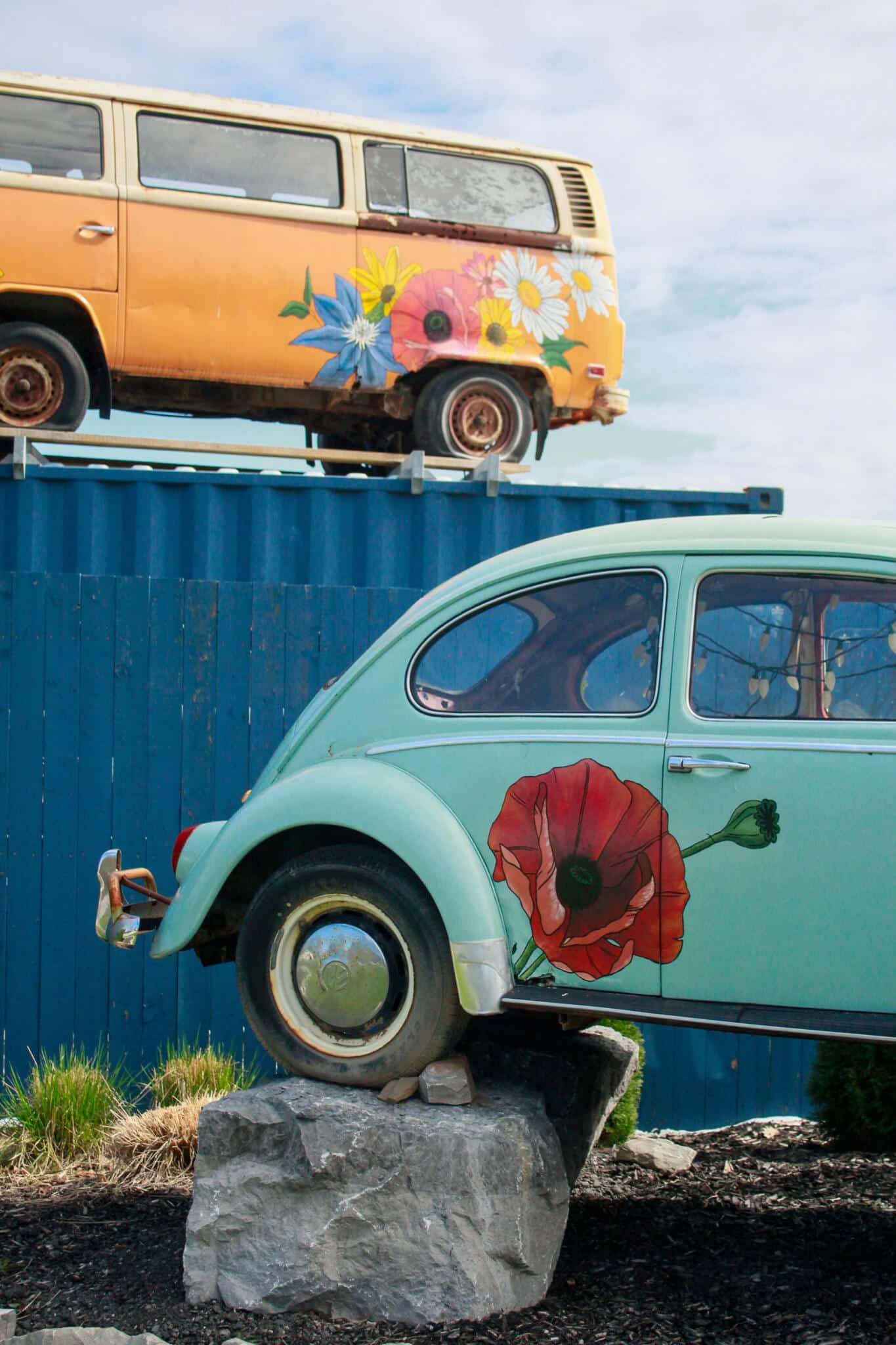 The Watering Can Vintage VW Beetle and Bus Display A vintage teal and blue Volkswagen Beetle displayed alongside an orange Volkswagen bus at The Watering Can Flower Market, both decorated with colourful floral motifs.