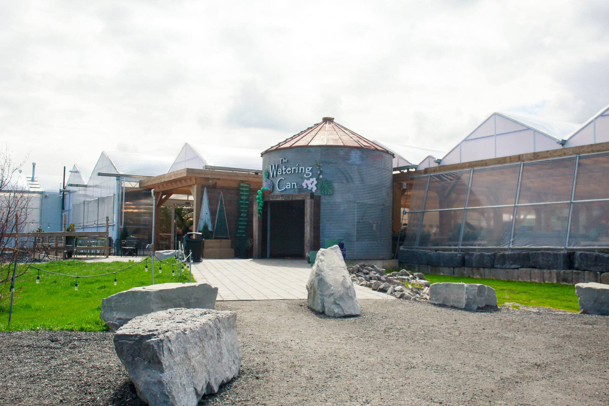 The Watering Can Grounds — Silo Buildings and Courtyard A wide-angle view of The Watering Can Flower Market grounds in Vineland, featuring the landmark silo buildings, a gravel pathway, and manicured landscaping under an overcast sky.