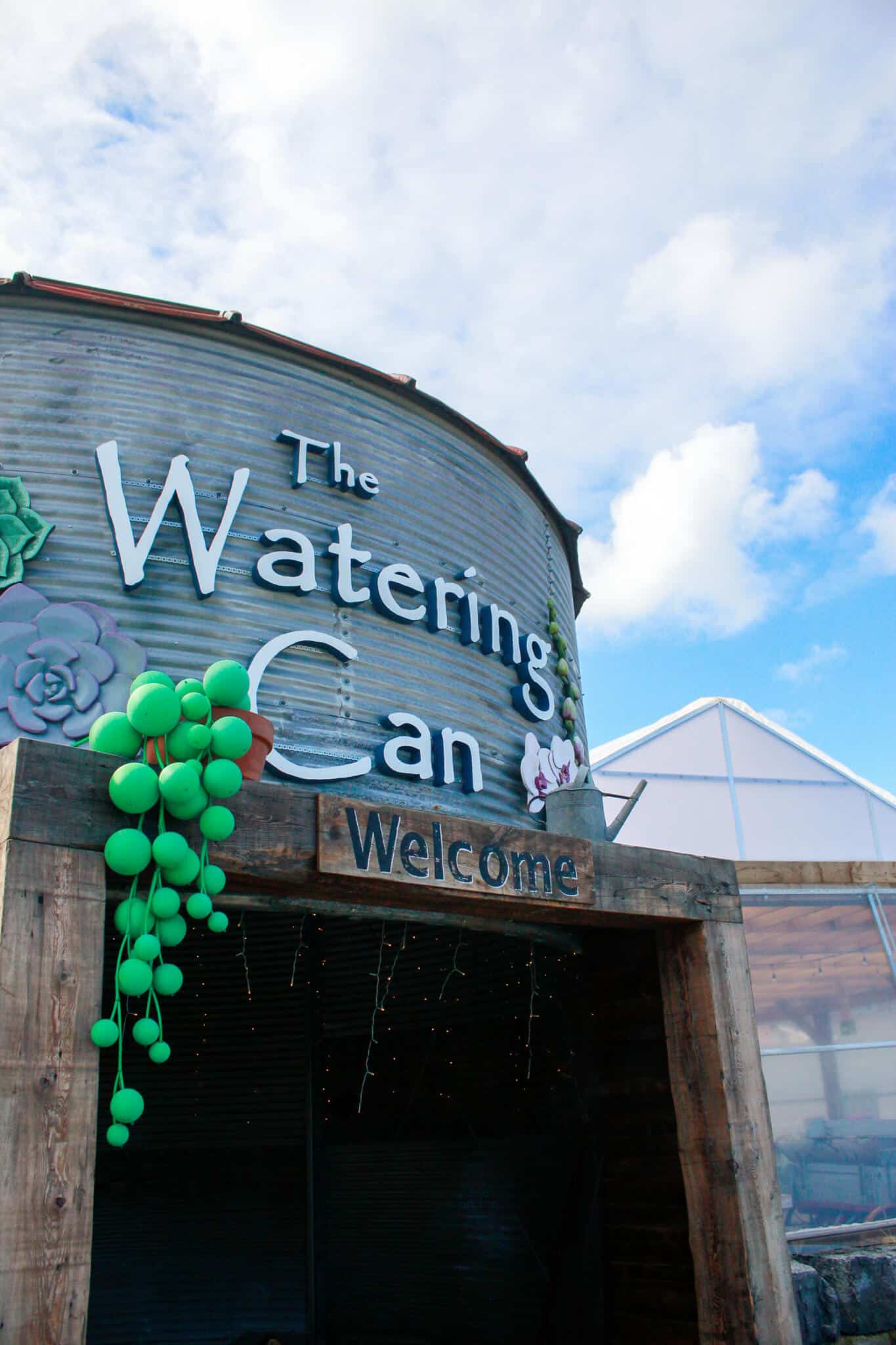 The Watering Can Flower Market Main Building Entrance The main entrance of The Watering Can Flower Market in Vineland, featuring the iconic wooden sign, green decorative balloons, and the signature silo building in the background.