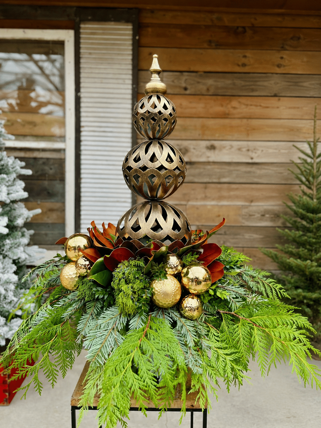 A winter porch urn topped with a tall bronze filigree finial, surrounded by evergreen boughs, magnolia leaves and gold ornaments.
