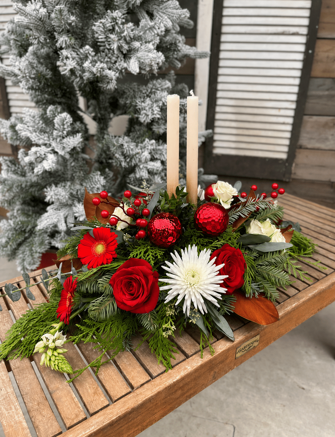 A festive fresh floral centrepiece with two ivory taper candles, red roses and gerberas, a white spider chrysanthemum, red ornaments and berries nestled in evergreens and eucalyptus, on a wooden bench.