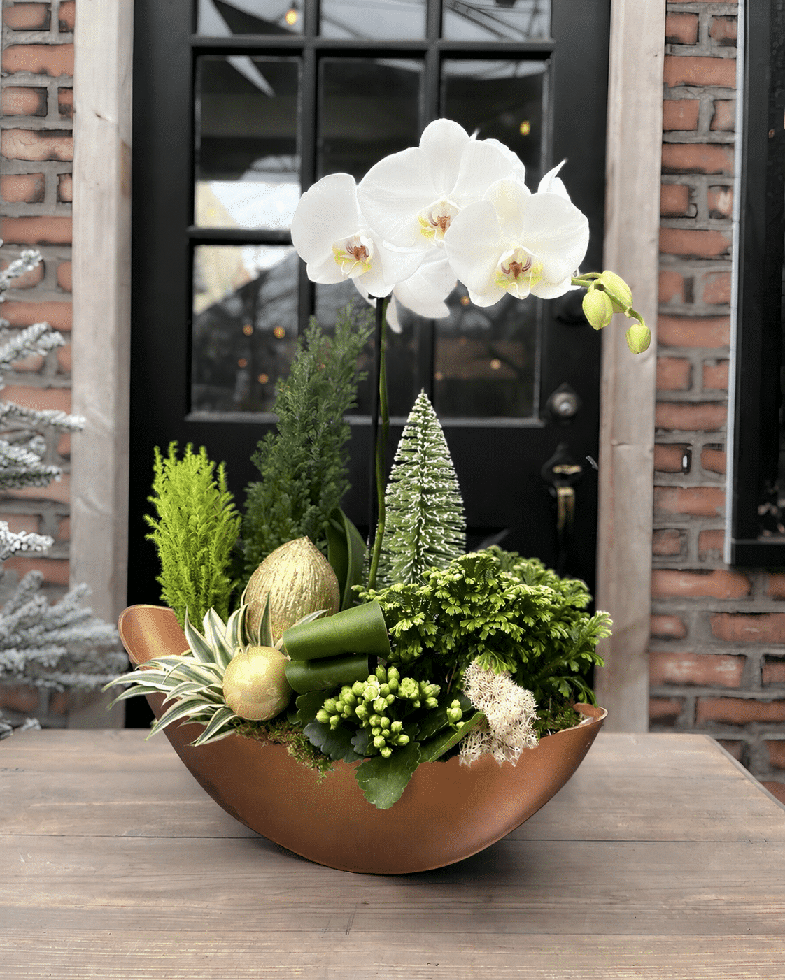 A holiday planted arrangement in a copper bowl featuring a white phalaenopsis orchid, fresh evergreen sprigs, a frosted bottlebrush tree, and a gold ornament.