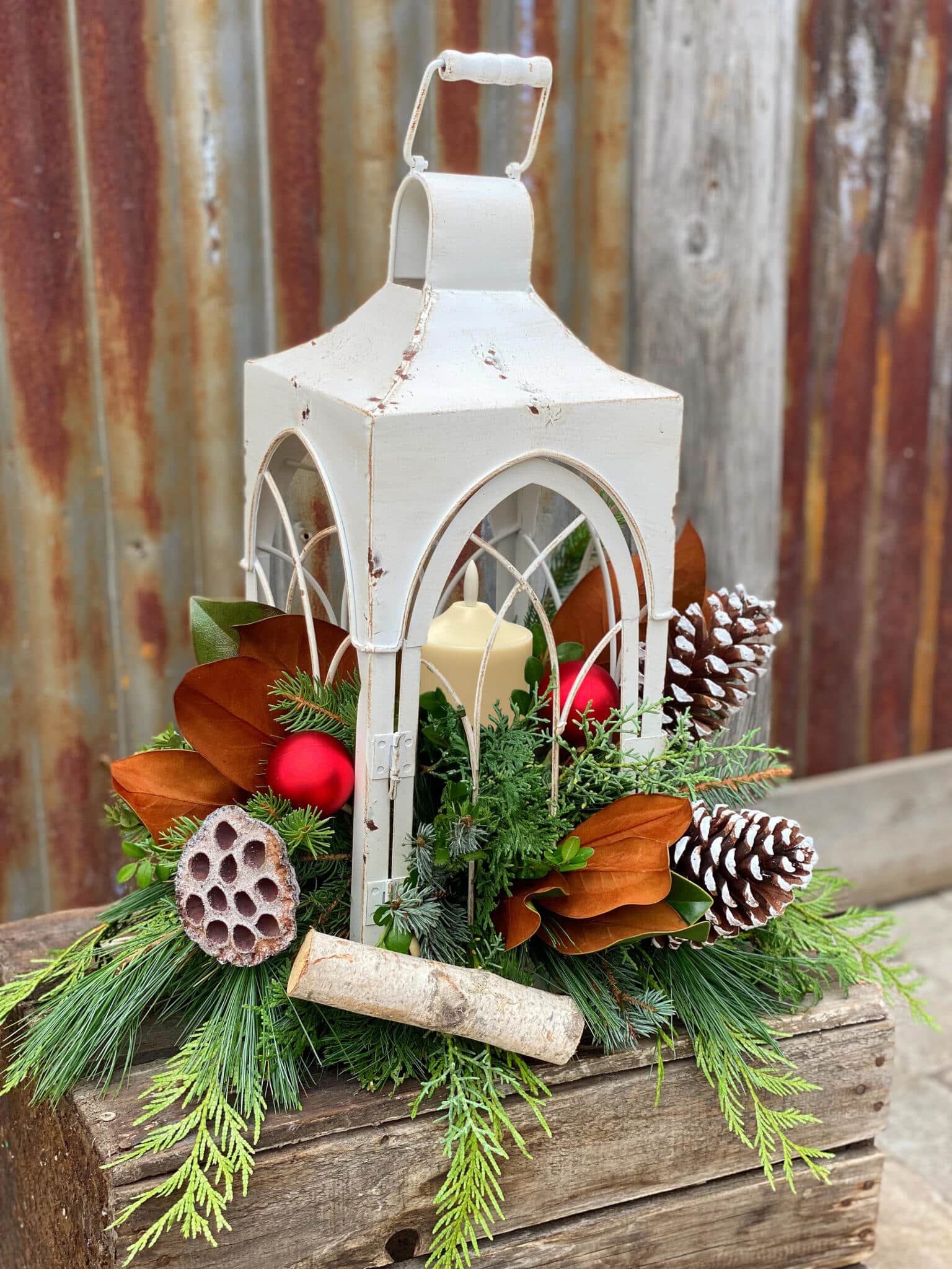 A distressed white metal lantern holding a candle, surrounded at its base by fresh evergreens, magnolia leaves, pinecones, red ornaments and a birch log, set on a rustic wood crate.
