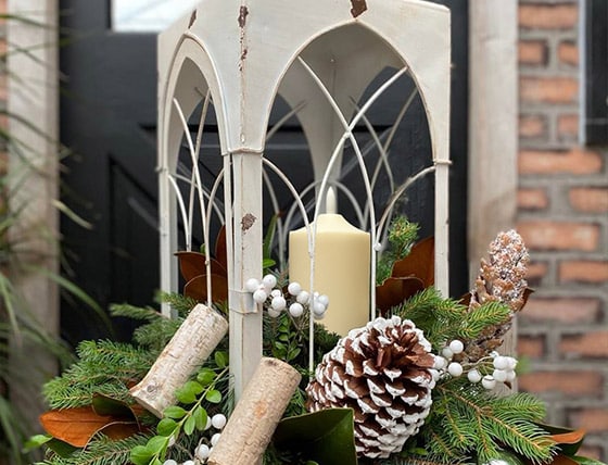 Cream metal lantern with arched windows and a pillar candle, surrounded by evergreens, birch logs and frosted pinecones.