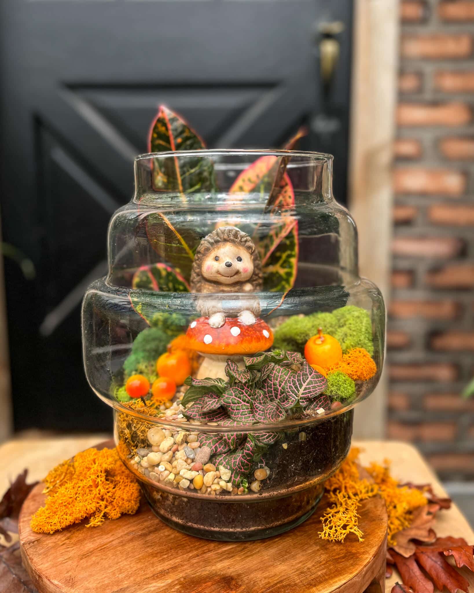 Glass terrarium with a hedgehog figurine on a red toadstool, tropical leaves, orange berries and moss, set on a wood slice.